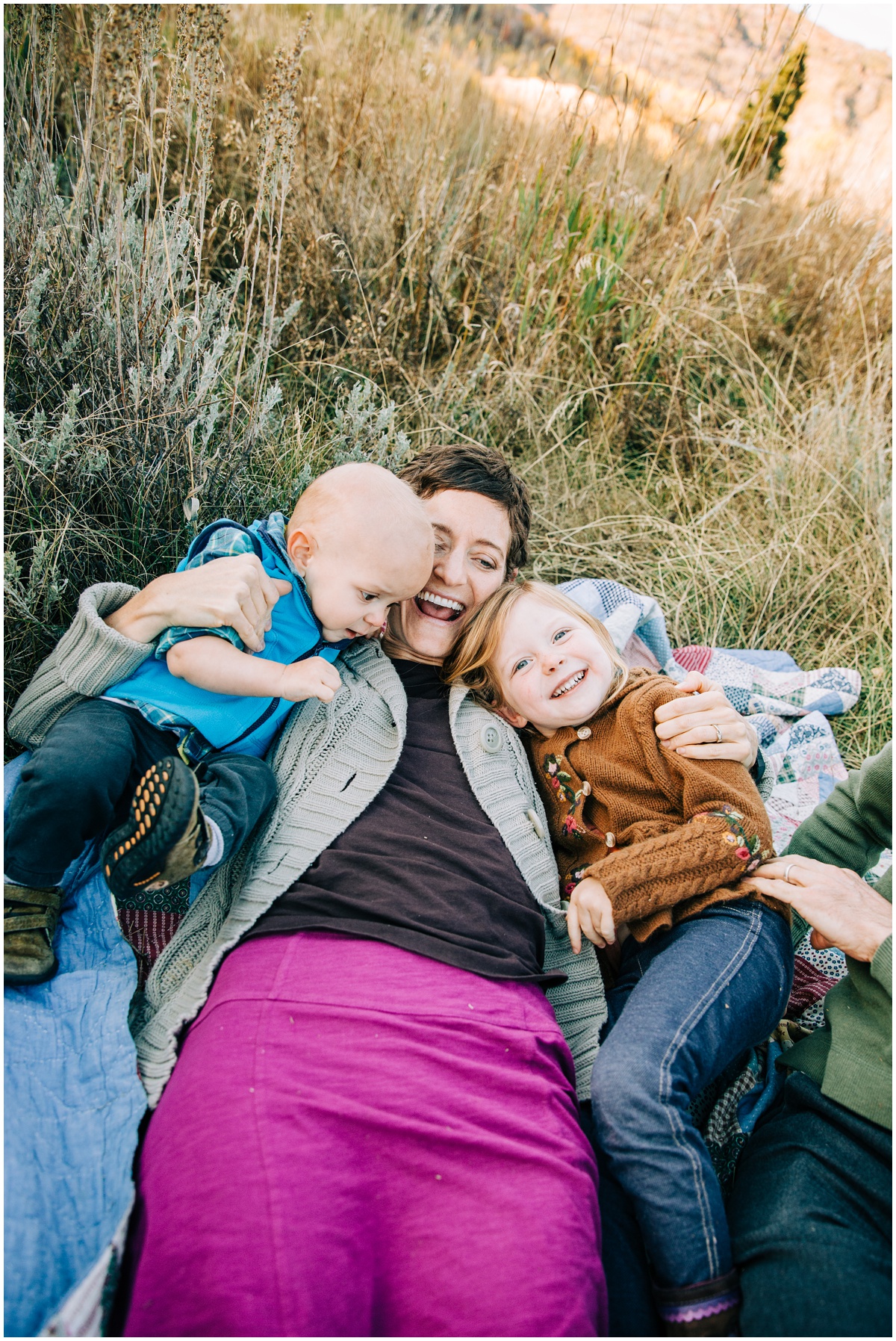 two kids playing with mom laying on blanket outside