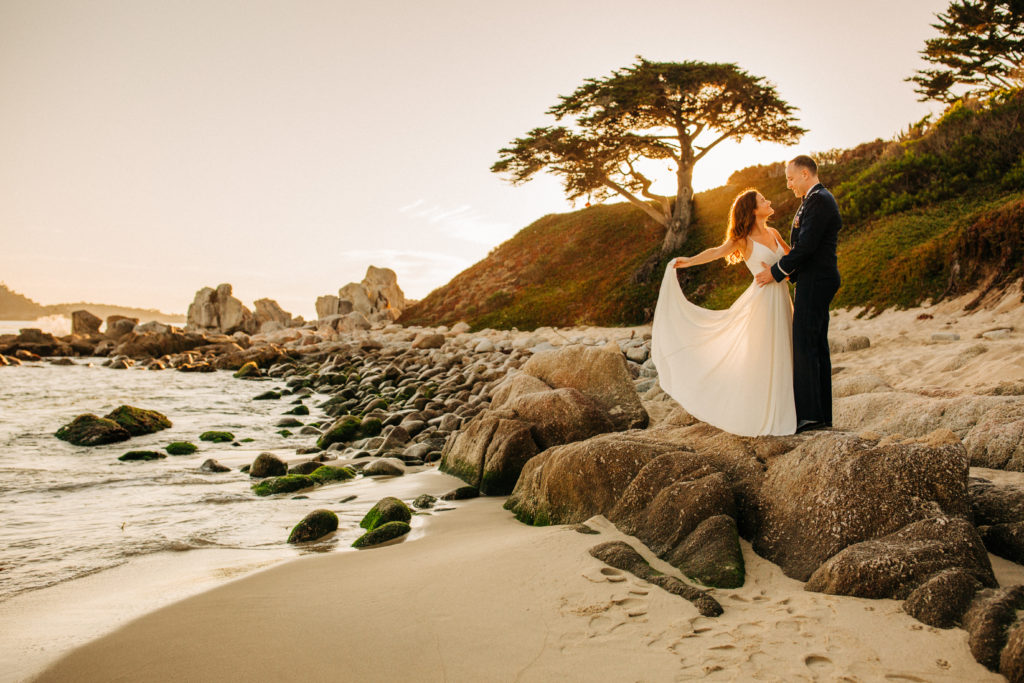 Bride and groom standing on coastal rocks at Carmel River Beach during their sunset elopement, with golden light illuminating her flowing dress and the iconic cypress tree in the background.
