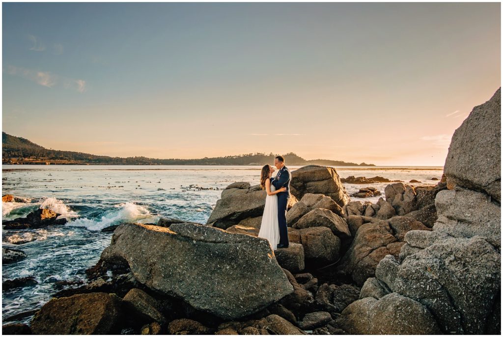 ouple embracing on large coastal rocks during their Carmel River Beach elopement, with waves crashing and golden California sunset light in the background