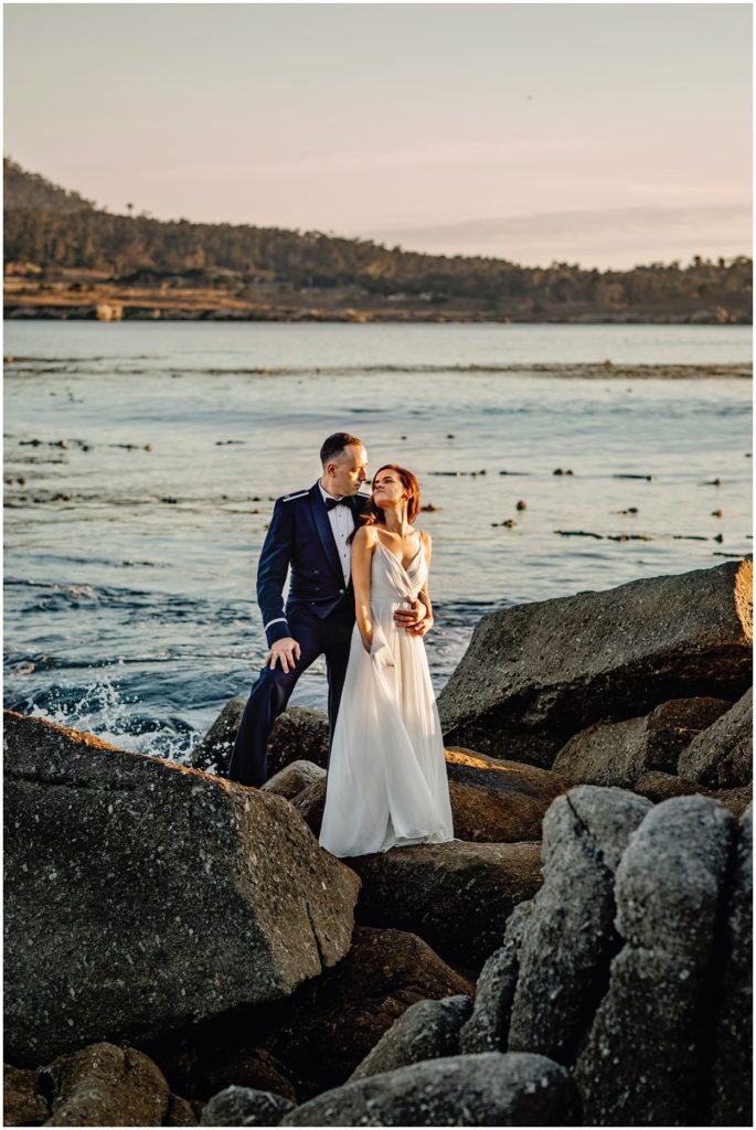 Bride and groom standing on the rocky shoreline at Carmel River Beach during their coastal elopement, with waves splashing behind them and soft golden California light