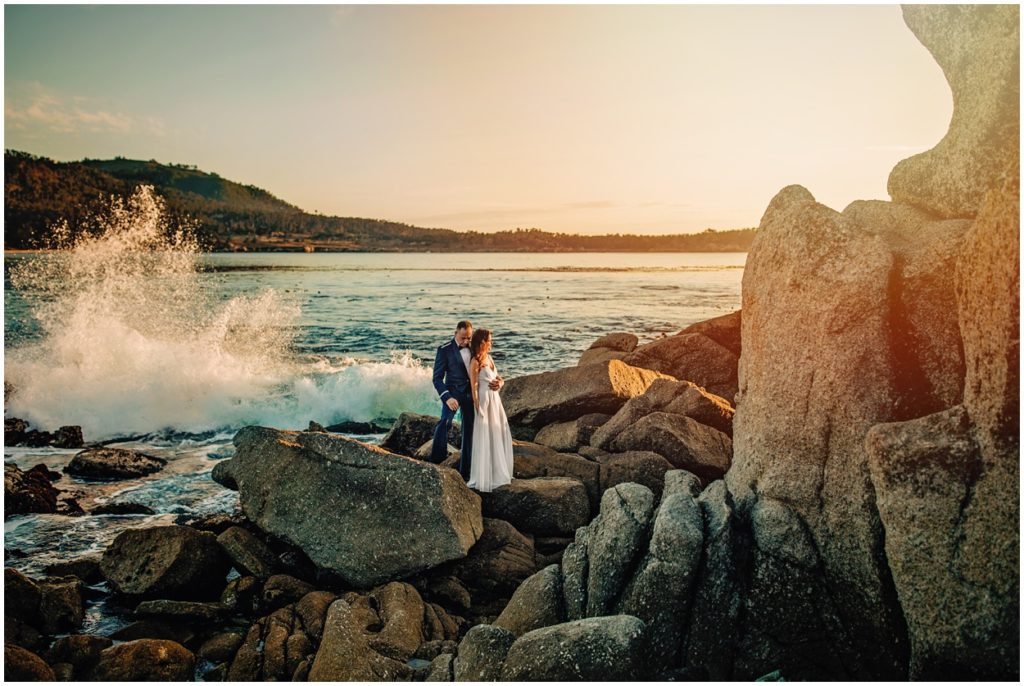 Bride and groom standing on dramatic coastal cliffs during their Carmel River Beach elopement, overlooking the ocean at golden hour with waves swirling below