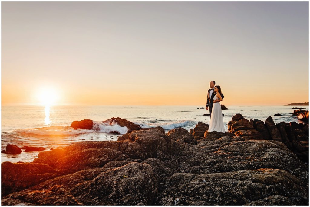 Bride and groom standing on rocky cliffs at Carmel River Beach, watching the sun set over the ocean during their intimate California coast elopement