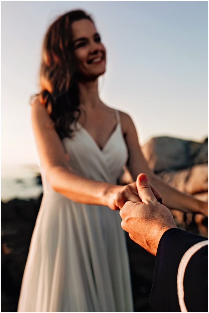 Bride smiling as she holds her groom’s hand during their Carmel River Beach elopement, captured in soft golden sunset light
