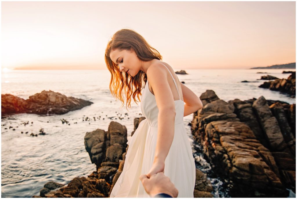Bride holding her groom’s hand while walking along the rocky shoreline during their Carmel River Beach elopement at sunset