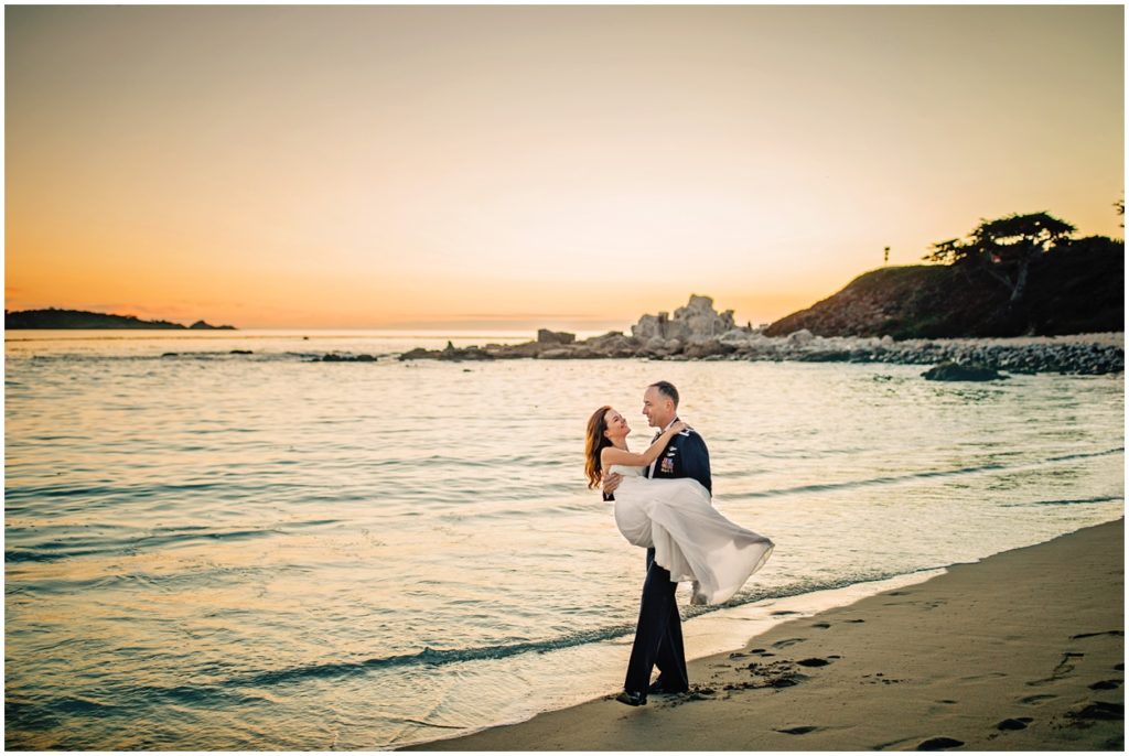 Groom carrying his bride along the shoreline during their Carmel River Beach elopement, with soft waves and a pastel sunset in the background