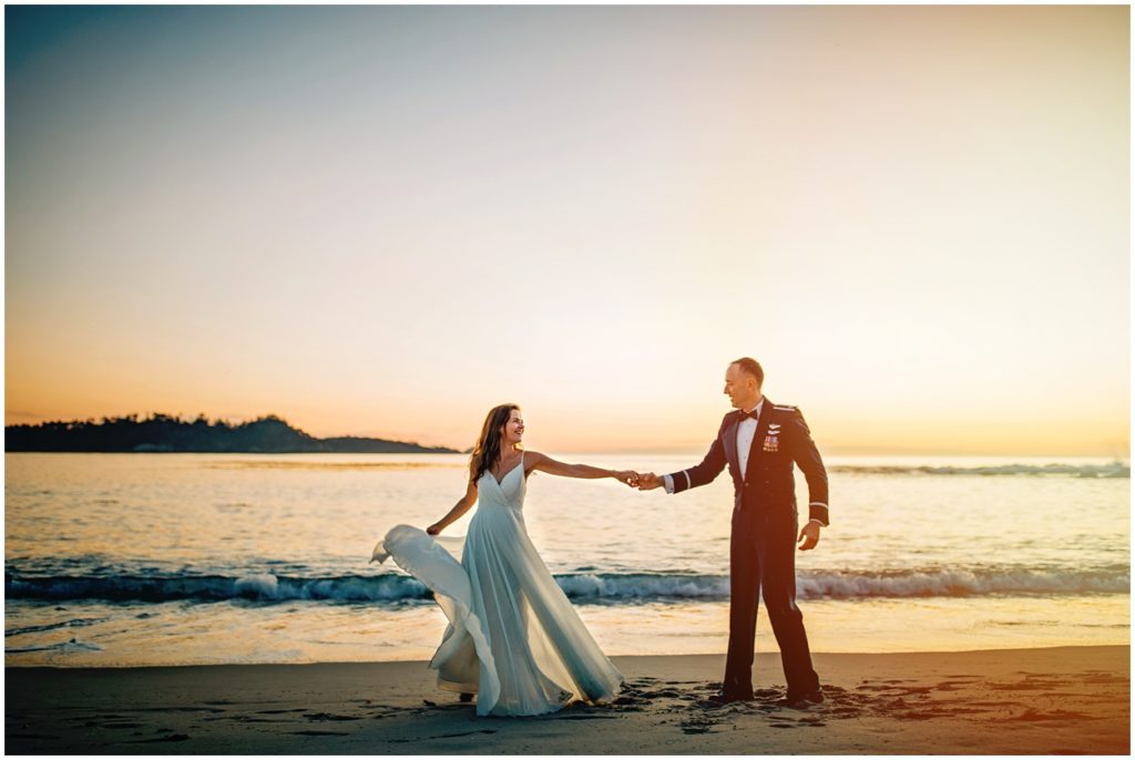 Bride and groom holding hands and dancing along the shoreline during their Carmel River Beach elopement at sunset, with soft waves and golden light behind them